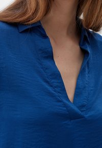 Blusa blu in cotone con scollatura a V profonda, realizzata in tessuto testurizzato, che presenta pieghe sottili e una vestibilità morbida attorno al colletto.