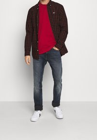 Rotes T-Shirt unter einem schwarz-roten karierten Hemd. Getragen mit blauen Jeans und weißen Turnschuhen mit schwarzen Akzenten.