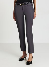 Pantalons ajustés gris en tissu lisse, présentant une coupe slim, des passants de ceinture et une boucle de ceinture en métal. Portés avec des chaussures à talons noires.