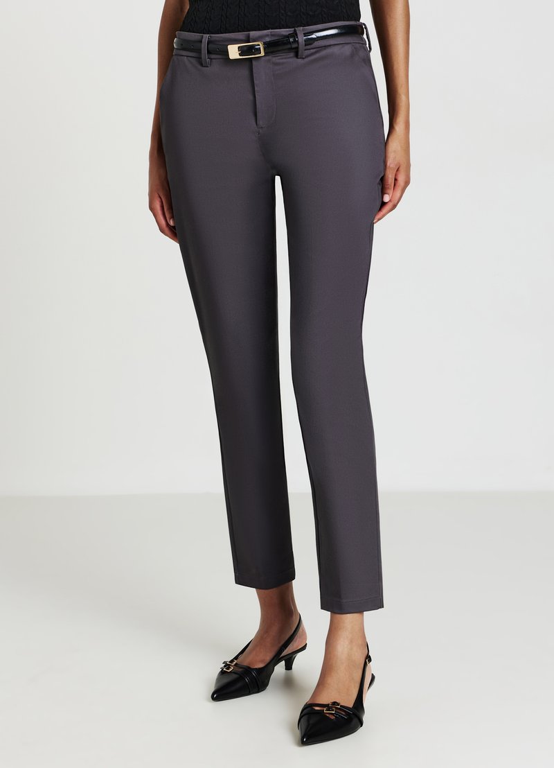 Pantalons ajustés gris en tissu lisse, présentant une coupe slim, des passants de ceinture et une boucle de ceinture en métal. Portés avec des chaussures à talons noires.