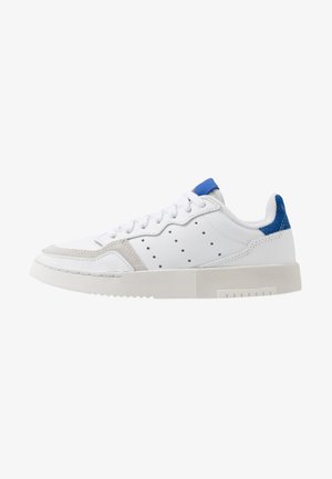 Witte lage sneaker met blauwe hieltab, geperforeerde zijdetails, witte veters en een dikke, getextureerde zool.
