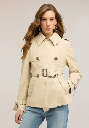 Frau mit langem, welligem Haar, trägt einen beigen Trenchcoat mit Gürtel und hellblauen Jeans, steht vor einem neutralen Hintergrund.