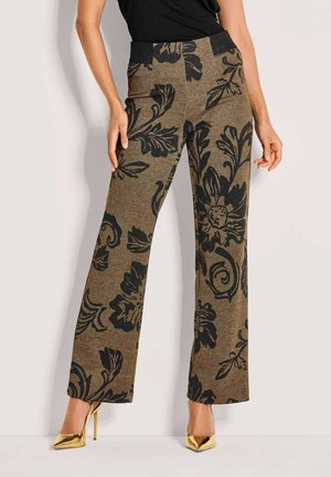 Frau in hoch taillierten braunen Hosen mit großen schwarzen Blumenmustern und goldenen spitzen High Heels vor einem schlichten Hintergrund.