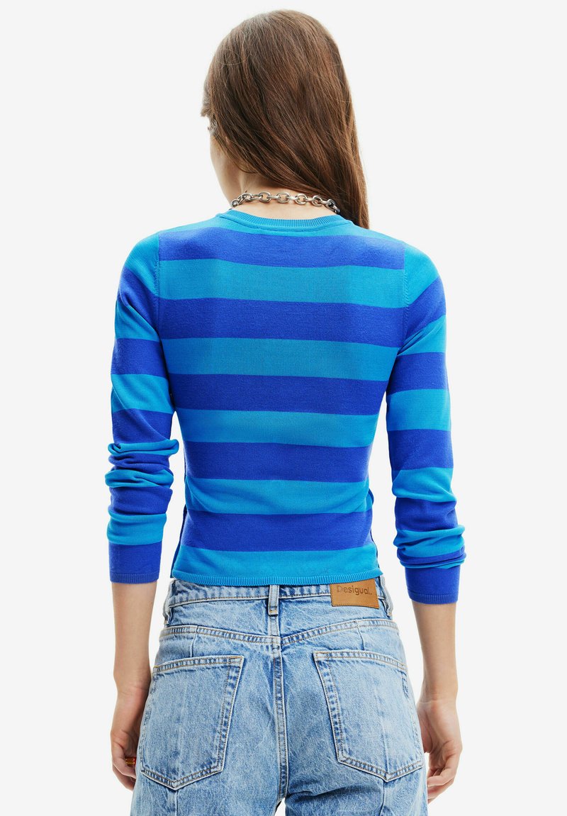 Mujer con cabello largo castaño, vistiendo un suéter a rayas azul y jeans de tiro alto azul claro, vista desde atrás con un fondo blanco.