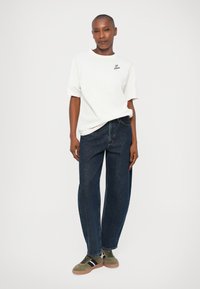 T-shirt bianca in cotone con maniche corte e logo sul petto, abbinata a jeans in denim blu scuro a vita alta e sneakers in suede verde.
