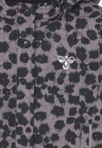 Jagten i leopardprint i sort og grå, med knaplukning, hætte og sølvbi-logo på brystet. Teksturstoffet har en blank finish.