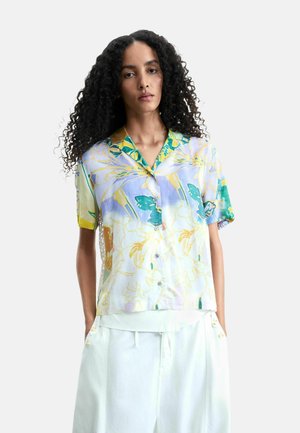 Kobieta o kręconych włosach, ubrana w pastelową kwiatową koszulę na krótki rękaw zapinaną na guziki oraz białe spodnie z wysokim stanem, stoi z rękami w kieszeniach.