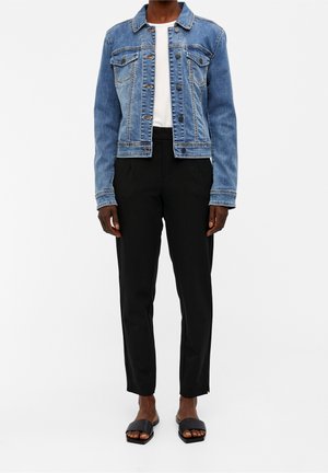 Person iført blå denimjakke, hvid top, sorte bukser og sorte åbne tå-sandaler, stående mod en ensfarvet baggrund.