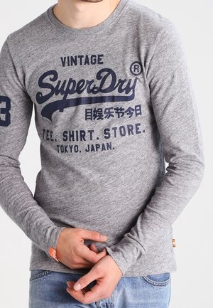 Homme portant une chemise grise à manches longues avec l'inscription « Vintage SuperDry » et Tokyo, Japon, ajustant les manches, jean bleu visible.
