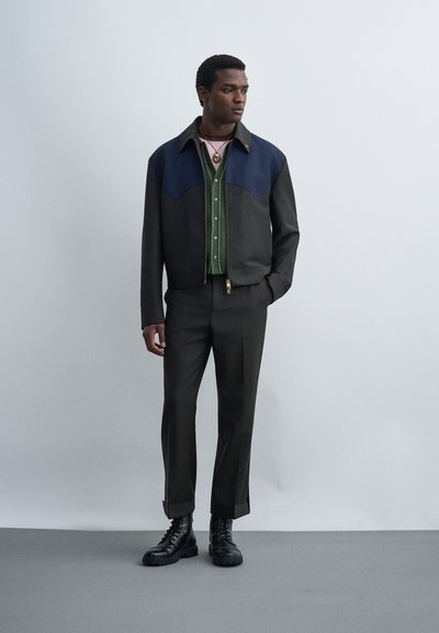 Άνδρας στέκεται, φοράει μαύρο και ναυτικό μπουφάν, πράσινο πουκάμισο με σχέδια, μαύρο παντελόνι, μαύρα μποτάκια, με τα χέρια στις τσέπες σε ουδέτερο γκρι φόντο.