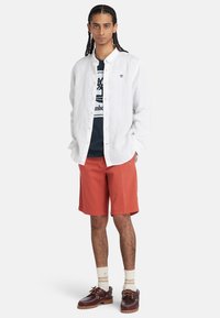 Giovane con capelli intrecciati che indossa una camicia bianca a maniche lunghe sopra una maglietta blu con logo, pantaloni corti color ruggine, calze color crema e scarpe da barca marrone scuro.