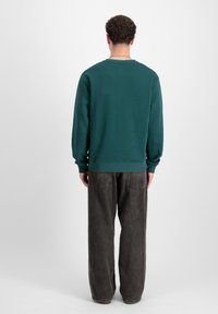 Sudadera de algodón verde con cuello redondo, puños acanalados y dobladillo. Llevada con pantalones oscuros y holgados, vista desde la parte de atrás.