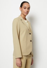 Blazer beige sartorial in un tessuto liscio, con colletto a rever, due tasche frontali e due bottoni marroni per la chiusura.