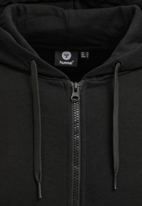 Svart huvtröja med dragkedja, tillverkad av mjukt material, med en framsluten dragkedja, dragsko vid huvan och en slät yta. Varumärkesetikett synlig på insidan.