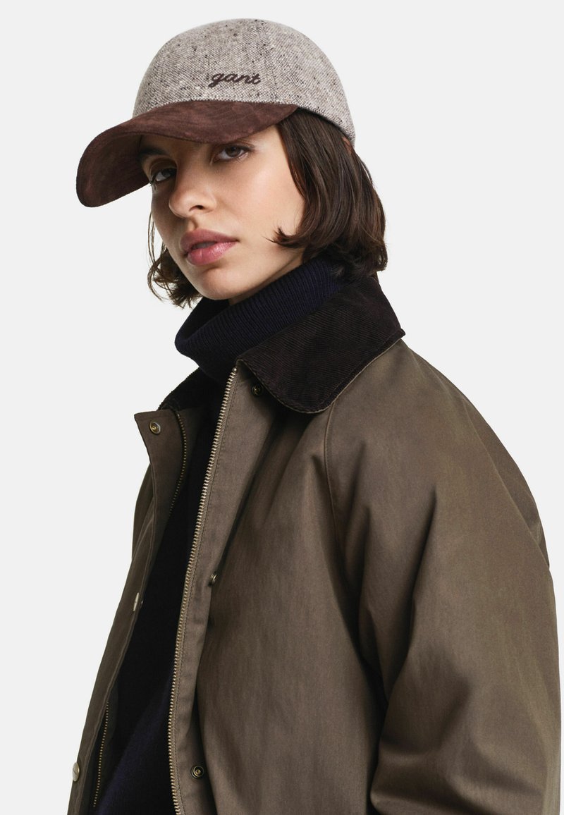 Brown cotton jacket with a zip closure, complemented by a dark collar; accompanied by a beige tweed cap featuring a brown visor and embroidered logo.