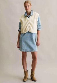 Denim dress with a light blue wash, featuring a scooped neckline and a frayed hem, layered with an off-white, cropped fleece vest.