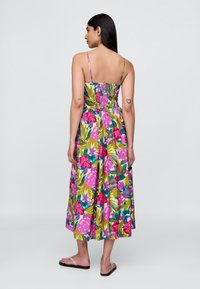 Vestido maxi floral com alças finas ajustáveis, padrão vibrante em rosa e verde, costas franzidas e saia fluida. Usado com sandálias rasas.