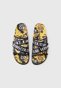 Sandálias pretas tipo slide com padrão barroco amarelo e texto branco a dizer "Versace Jeans Couture." Possuem tiras largas para suporte.