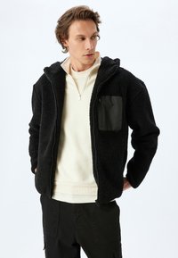 Chaqueta de forro polar negra con capucha, cierre de cremallera y un exterior texturizado. Cuenta con un bolsillo frontal negro y se lleva sobre un sudadera de color claro.