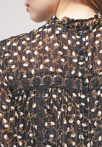 Blouse à fleurs noire avec des fleurs blanches et moutarde sur un tissu transparent, dotée d'un col à volants, d'une bordure en dentelle noire et d'une fermeture à boutons dans le dos.