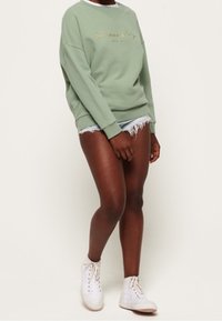 Jasnozielona bawełniana bluza z okrągłym dekoltem i złotym zdobieniem w formie pisma, zestawiona z postrzępionymi szortami jeansowymi i białymi trampkami.