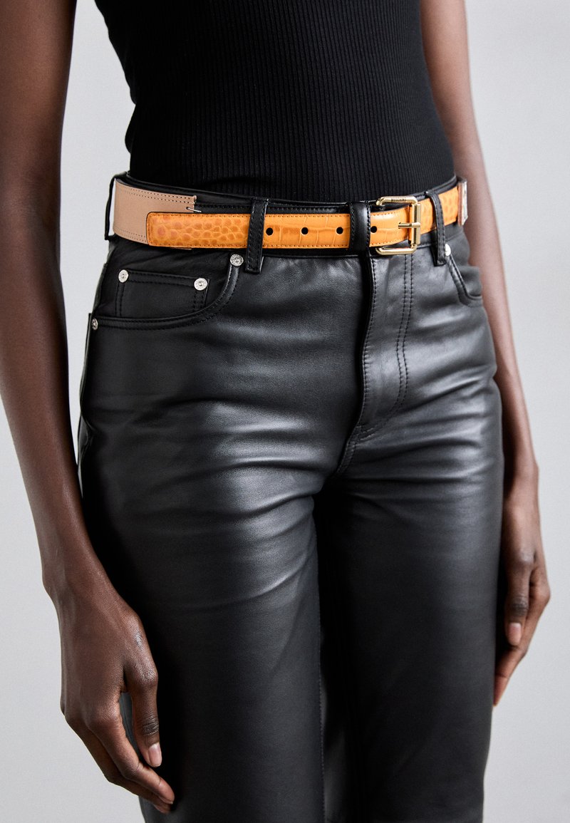 Pantaloni din piele neagră, asortați cu un curea bicoloră cu un model de crocodil în nuanțe de portocaliu și bej, fixată cu o cataramă metallică.