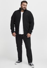 Giacca in pile nera con colletto alto, chiusura con zip e maniche lunghe; indossata sopra una maglietta bianca e abbinata a pantaloni neri e sneakers.
