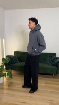 Hanorac gri cu glugă mare, buzunar frontal și manșete tricotate, asortat cu pantaloni negri cu picioare largi și pantofi negri. Canapea verde în fundal.