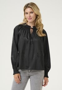 Blusa nera con motivo floreale sottile, scollatura arricciata con chiusura a legaccio, maniche lunghe con polsini aderenti, tessuto testurizzato, vestibilità rilassata.