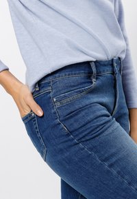 Personne portant un jean bleu et un haut à manches longues bleu clair, main placée de manière décontractée dans la poche avant sur un fond uni.