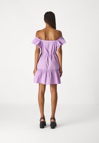 Frau mit geflochtenem Haar, die ein lilafarbenes schulterfreies Kleid und schwarze Blockabsatzsandalen trägt, steht von hinten vor einem schlichten weißen Hintergrund.