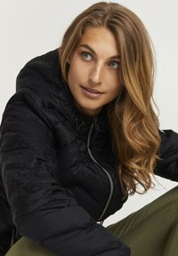 Juodas pūkinis švarkas su kapišonu, užsegamas užtrauktuku ir turintis šniūrą. Lygus paviršius ir siuvinėjimo dizainas. Modelis vilki alyvų žalius kelnes.