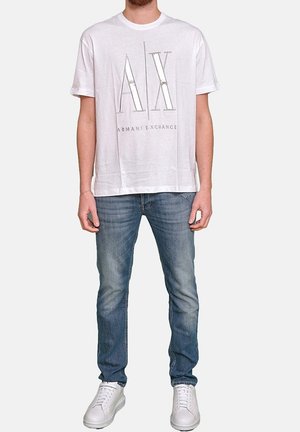 Hvid bomuld t-shirt med stort grafisk logo "AX" og "ARMANI EXCHANGE" trykt i grå. Båret med blå jeans og hvide sneakers.
