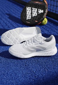 Chaussures de sport blanches sur une surface de court bleue avec une raquette de padel noire et une balle jaune près d'un filet.
