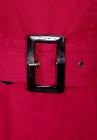 Ljusrosa bälte gjort av tyg, med en stor rektangulär svart plastspänne med en guldmetskrok. Strukturerad yta med sömnadsdetaljer.