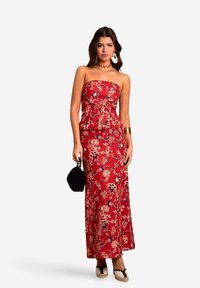 Rode bloemen maxi-jurk met een strapless, peplum-top. De stof heeft een bloemenpatroon; het model houdt een kleine zwarte tas vast en draagt espadrilles.