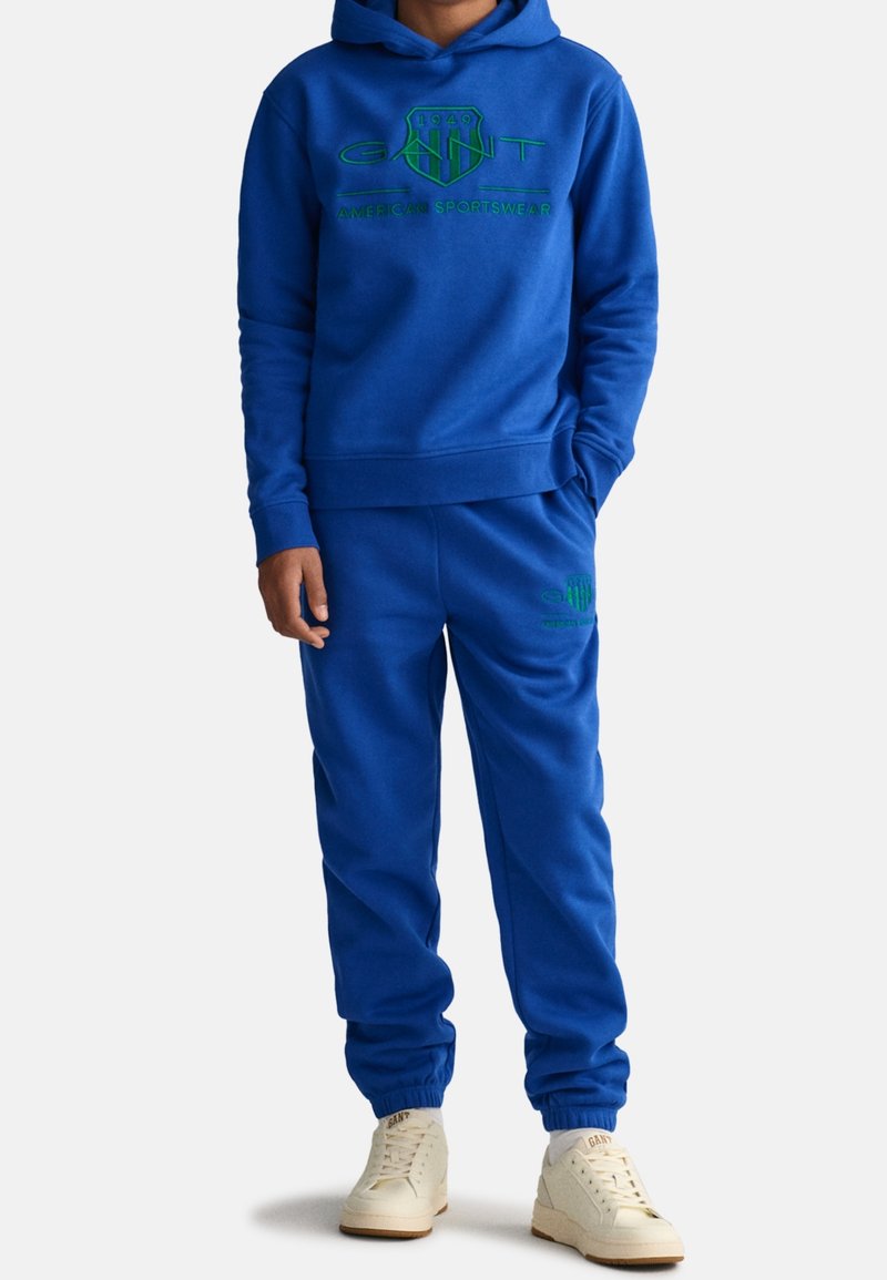 Blå sweatshirt och matchande joggers i mjukt material. Har grön logotext på bröstet och på vänster ben. Vita sneakers.