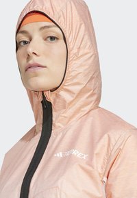 Veste Terrex rose clair avec capuche à cordon de serrage et zip sur toute la longueur. Tissu texturé avec fermeture éclair et logo noirs contrastants.