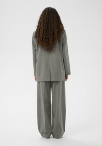 Femeie cu păr lung și creț, îmbrăcată într-un costum gri cu dungi, stând cu spatele într-un fundal alb simplu.