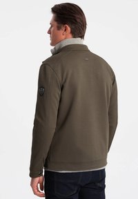 Giacca verde oliva con finitura testurizzata, colletto alto grigio e emblema nero sulla manica, caratterizzata da un design aderente e cuciture sottili.