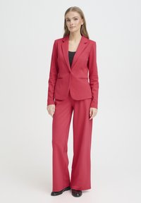 Blazer rouge cintré associé à un pantalon à jambes larges. Tissu lisse, fermeture à un bouton sur le blazer, et un design classique de col à revers. Haut noir en dessous.