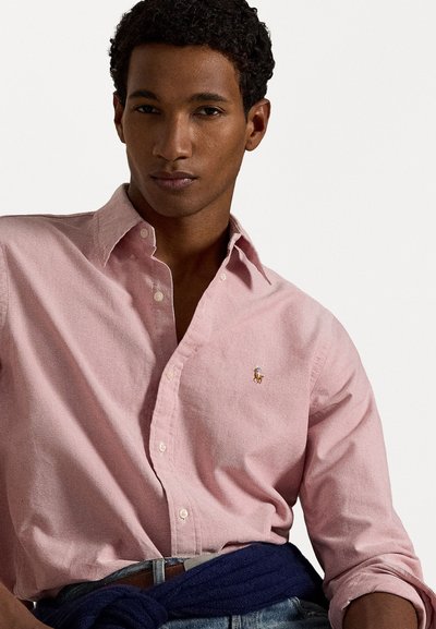 Ljust rosa skjorta med knappar, tillverkad av mjukt tyg, med en liten broderad logotyp på bröstet och en krage, stylad med uppvikta ärmar.