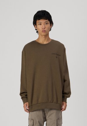 Bruine sweatshirt met lange mouwen, een ribbelboord bij de crewneck en manchetten. Heeft een klein bedrukt ontwerp op de linkerkant van de borst. Zacht, gestructureerd materiaal.