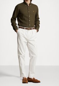 Muž v olivově zelené košili na knoflíky, bílých plisovaných kalhotách s hnědým páskem a hnědých semišových mokasínách s třásněmi, stojící s rukama v kapsách.