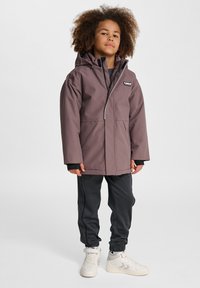 Wasserabweisende mauvefarbene Jacke mit Kapuze, Frontreißverschluss und zwei großen Taschen. Kombiniert mit schwarzen Jogginghosen und weißen Turnschuhen.