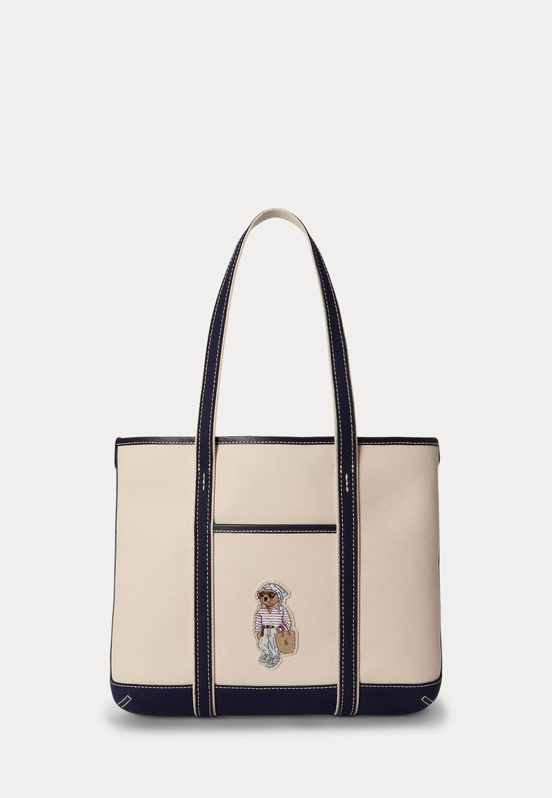 Beige totebag med mørkeblå kant og håndtak, med en liten brodert bjørn som har på seg en stripete skjorte og hatt.