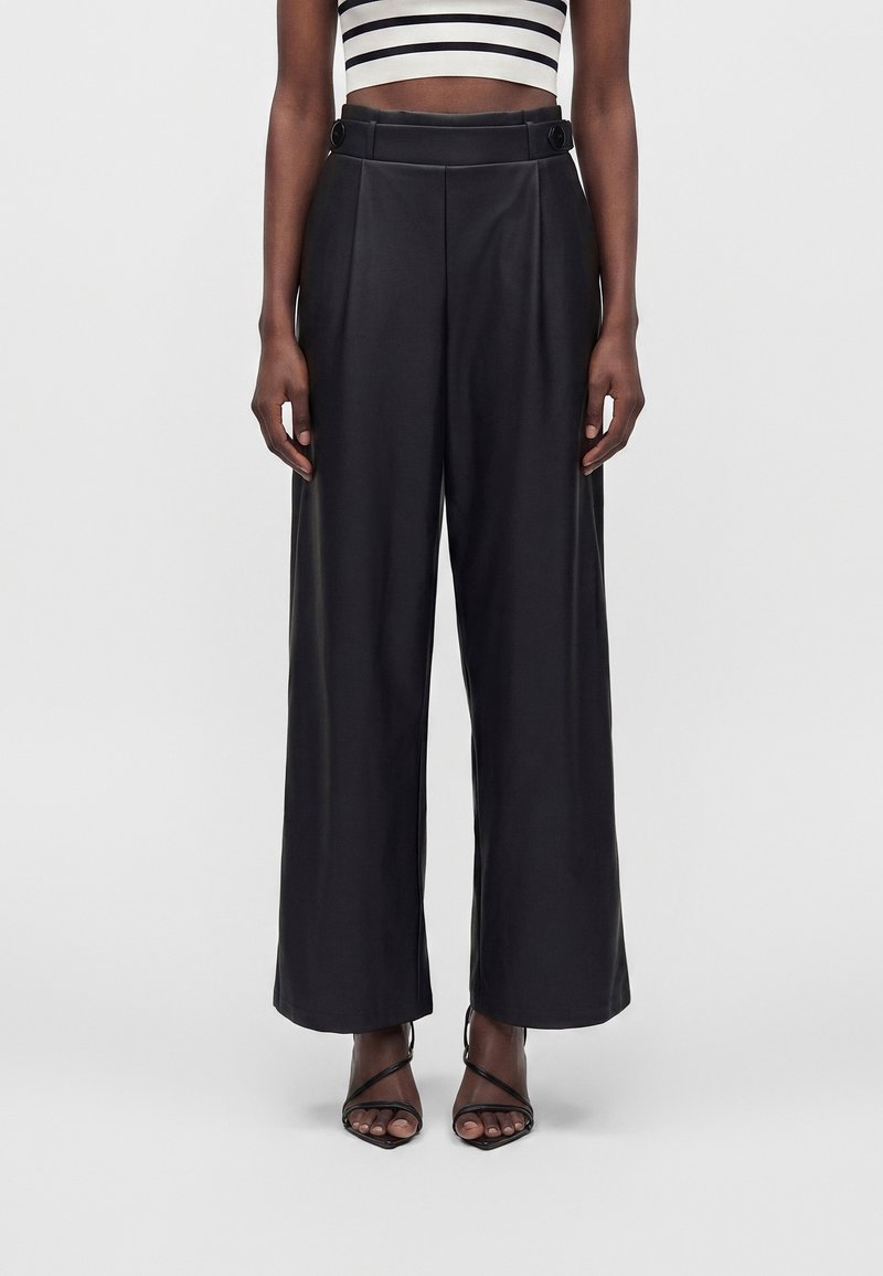 Femme portant un pantalon noir taille haute à jambes larges avec des détails de boutons, associé à des talons noirs à brides ouvertes et un top court rayé noir et blanc.