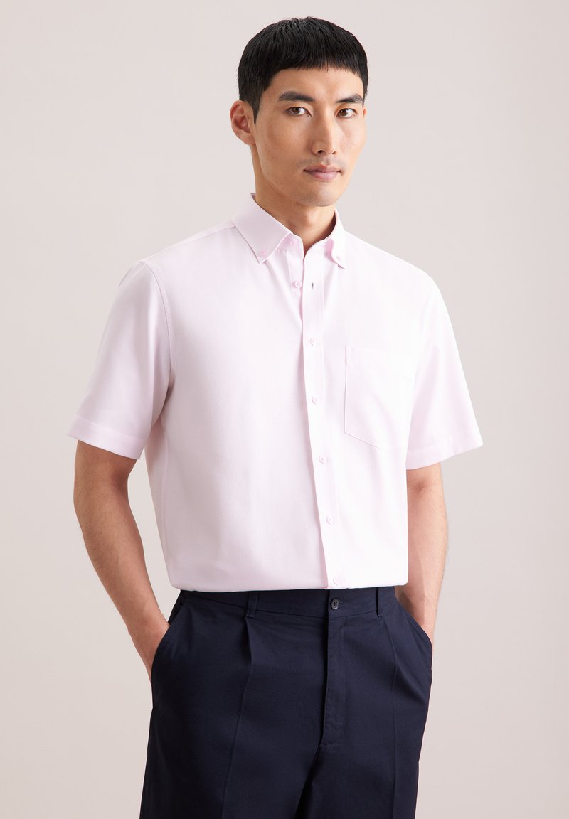 Homme portant une chemise à manches courtes rose clair avec une poche poitrine et un pantalon bleu marine foncé, les mains dans les poches, fond neutre.