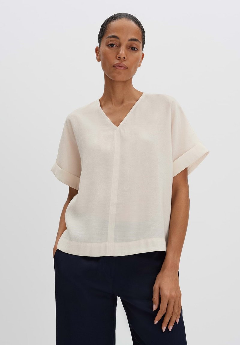 Blusa crema a maniche corte con scollo a V, caratterizzata da una texture liscia e una sottile cucitura frontale, abbinata a pantaloni blu scuro.