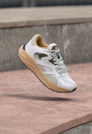 Baskets athlétiques blanches et beiges avec des lacets gris, suspendues en l'air au-dessus de marches en béton texturé.